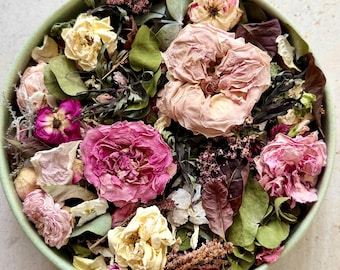 May include: A round, light green bowl filled with a collection of dried flowers and foliage. The arrangement includes various shades of pink, cream, and white roses, along with dark purple and green dried leaves and small textured botanical sprigs.