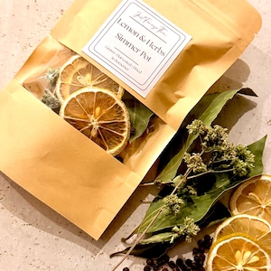 May include: A tan pouch with a clear window containing dried lemon slices and herbs. The label reads Just Forage This, Lemon & Herbs Simmer Pot, Grown, Harvested, Dried in Somerset. Loose dried lemon slices, green leaves, and peppercorns are nearby.