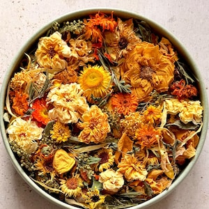 May include: A round, light green bowl filled with a dense collection of dried flowers in various shades of yellow, orange, and gold. The assortment includes dried marigolds, daisies, and other small blossoms, creating a textured, vibrant display.