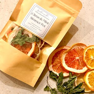 May include: A tan paper pouch containing dried citrus slices and mint leaves with a label reading Citrus & Mint Simmer Pot, Grown-Harvested-Dried in Somerset. Beside the pouch are loose dried grapefruit slices, orange slices, and green mint leaves.