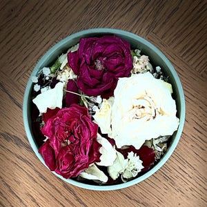 May include: A round, light blue bowl filled with dried flowers. The arrangement includes deep red and cream-colored roses, along with small white flowers and petals. The bowl sits on a wooden surface, creating a natural and rustic aesthetic.