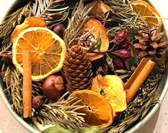 Op de afbeelding: Een ronde, lichtgroene kom gevuld met een potpourri-mix van gedroogde sinaasappelschijfjes, kaneelstokjes, dennenappels, gedroogde appelschijfjes, rozenbottels, laurierblaadjes en groenblijvende takjes met natuurlijke texturen.