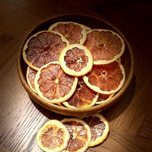 May include: A wooden bowl containing dried orange slices. The slices are a mix of brown and orange, with visible segments and a circular shape. The bowl rests on a wooden surface, creating a warm, natural aesthetic.