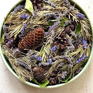Op de afbeelding: Een ronde, lichtgroene kom gevuld met een mengsel van gedroogde lavendeltakjes, bruine dennenappels, groene laurierblaadjes en gedroogde dennennaalden. Het arrangement bevat een verscheidenheid aan texturen en natuurlijke aardetinten, waaronder paars, bruin en groen.