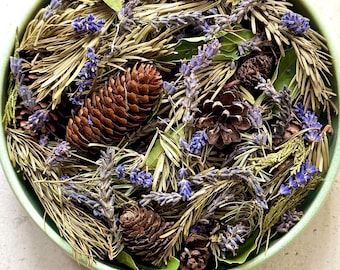 May include: A round, light green bowl filled with a mixture of dried lavender sprigs, brown pinecones, green bay leaves, and dried pine needles. The arrangement features a variety of textures and natural earth tones, including purple, brown, and green.