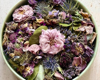 May include: A round, light green ceramic bowl filled with a collection of dried flowers and botanicals in shades of purple, pink, and green. The arrangement includes dried roses, lavender sprigs, thistle heads, and various green leaves.