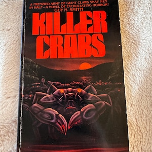 May include: A mass-market paperback book titled Killer Crabs by Guy N. Smith. The black cover features a large, dark purple crab with glowing orange eyes in the foreground, a swarm of smaller crabs behind it, and a red sun setting over a dark landscape.