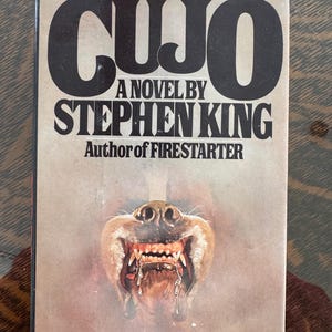 May include: Vintage paperback book titled "CUJO" by Stephen King. The cover features a menacing dog's face with bared teeth and drool, set against a muted background. The title and author's name are in bold, black lettering.