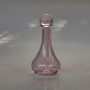 May include: A small, translucent light pink glass bottle with a teardrop-shaped body and a narrow neck. The bottle is topped with a clear, spherical glass stopper that sits securely in the opening, creating a smooth and continuous silhouette.