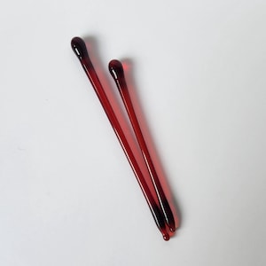 May include: Two red glass stir sticks with rounded tips. The sticks are slender and have a dark red color at the tips, gradually fading to a lighter red. They are positioned diagonally against a white background.