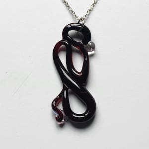 Puede incluir: Un collar con un colgante de vidrio negro y una cadena de plata. El colgante tiene forma de serpiente con una pequeña cuenta de vidrio transparente en la parte superior.