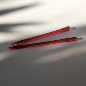 Peut inclure: Deux longs et fins bâtonnets en verre rouge sont disposés en diagonale sur un fond blanc. Les bâtonnets ont des extrémités arrondies et sont légèrement effilés. L'éclairage crée des ombres, mettant en valeur les formes et les textures du verre.