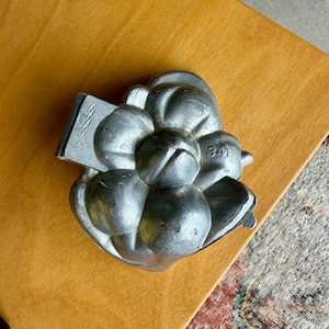May include: A silver-colored metal mold in the shape of a flower, with multiple rounded petals and a rectangular handle. The number "348" is visible on the side of the mold. The mold is placed on a light brown wooden surface.