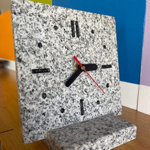 Puede incluir: Un reloj de escritorio cuadrado de granito con manecillas negras y un segundero rojo. La esfera del reloj tiene marcadores negros y está sostenida por una base rectangular de granito. La esfera y la base del reloj tienen un patrón moteado gris y blanco.