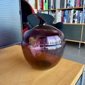 May include: A decorative, dark purple glass jar with a matching lid and a curved handle. The jar is round with a bulbous shape and sits on a wooden surface. The lid has a pointed, horn-like handle. The jar reflects the surrounding environment.