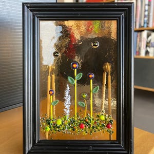 Può includere: Un'opera d'arte in vetro incorniciata con una scena di giardino. L'opera include steli gialli, foglie verdi e fiori colorati con accenti rossi e gialli. La cornice nera ha un supporto bianco.
