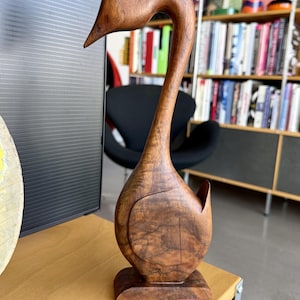 May include: A handcrafted wooden swan sculpture with a long, curved neck and detailed feather carving. The swan is made of dark brown wood and stands on a rectangular base. The sculpture is displayed on a light-colored wooden surface.