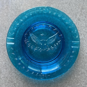 May include: A round, turquoise glass ashtray with a detailed embossed design. The center features an eagle with outstretched wings, surrounded by a wreath. The rim has a decorative pattern and two cigarette rests.
