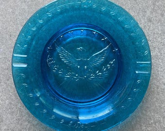 May include: A round, turquoise glass ashtray with a detailed embossed design. The center features an eagle with outstretched wings, surrounded by a wreath. The rim has a decorative pattern and two cigarette rests.