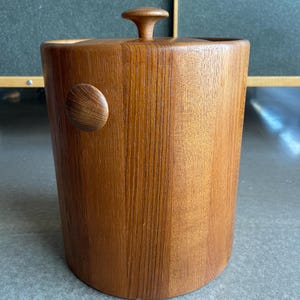 Puede incluir: Una cubitera cilíndrica de madera con tapa y asa lateral. La cubitera es de madera marrón claro con un patrón de vetas visible. La tapa tiene un pequeño asa redondeada. El lateral tiene un asa circular de madera.