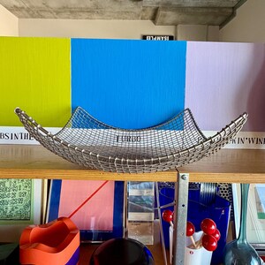 May include: A square, silver-toned wire mesh basket with curved, upward-sweeping edges. The basket features a grid pattern and is positioned centrally. The metal wire construction creates a semi-transparent, open-weave appearance with a metallic finish.