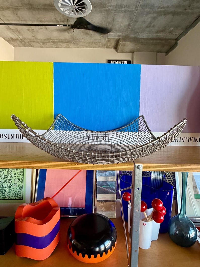 May include: A square, silver-toned wire mesh basket with curved, upward-sweeping edges. The basket features a grid pattern and is positioned centrally. The metal wire construction creates a semi-transparent, open-weave appearance with a metallic finish.