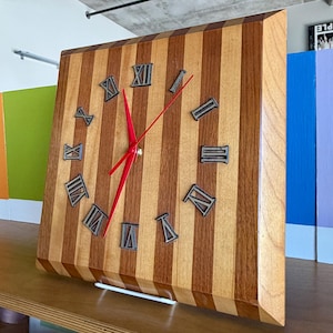 May include: A square wooden clock with a striped pattern in light and dark brown. The clock face features Roman numerals and red hands. The clock is displayed on a wooden shelf.