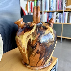 May include: A ceramic vessel with a rounded body, a central arched handle, and two small spouts on the upper shoulders. The surface features a marbled glaze pattern with swirling streaks of dark brown, tan, and cream colors across the exterior.