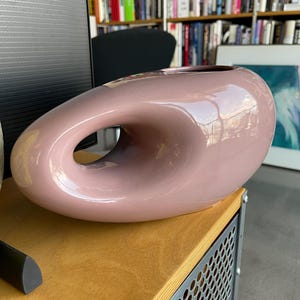 May include: A dusty rose-colored ceramic vase with a unique, organic shape. The vase features a large, open loop in the center, creating a distinctive design. The glossy finish reflects light, highlighting the smooth curves of the vessel. A modern decorative piece.