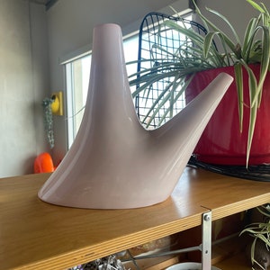 May include: A light pink ceramic vase with a unique, abstract design. The vase has a wide base and two spouts that curve upwards.