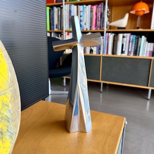 May include: A silver-colored cross sculpture with a unique, modern design. The cross has a flared base and a textured surface, standing on a wooden surface. The background includes a bookshelf filled with books and decorative items.