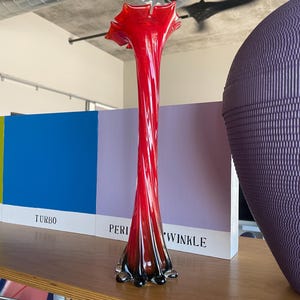 May include: A red glass vase with a twisted design and a black base. The vase is sitting on a wooden shelf with a blue and purple book behind it.