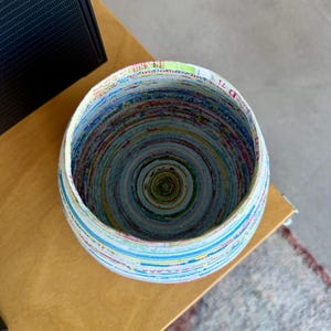 May include: A round bowl made from tightly rolled paper, showcasing a spiral pattern of colorful stripes. The bowl's rim is white, and the interior reveals a concentric design of various hues. The bowl sits on a light brown surface.