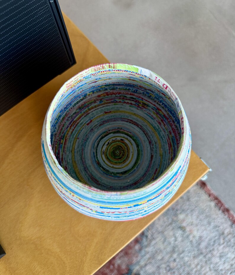 May include: A round bowl made from tightly rolled paper, showcasing a spiral pattern of colorful stripes. The bowl's rim is white, and the interior reveals a concentric design of various hues. The bowl sits on a light brown surface.