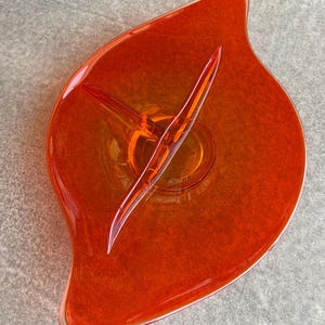 May include: A vibrant orange glass serving dish with a leaf-like shape. The dish features a central, raised section and a clear, pointed divider. The glass has a speckled texture, and the dish is set against a neutral background.