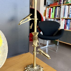 Op de afbeelding: Een messing standaard met een verticale stang met een bolvormige bovenkant, een decoratief handvormig element in het midden en twee verstelbare metalen clips aan de zijkanten. De voet is cirkelvormig met een gedetailleerd, geschulpt, bladachtig ontwerp.