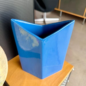 May include: A vibrant blue ceramic vase with a unique angular design. The vase has a textured, vertically ribbed surface and a glossy finish. The opening is shaped like a V, and the vase is sitting on a wooden surface.