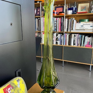 Könnte beinhalten: Eine hohe, grüne Glasvase mit einer einzigartigen, länglichen Form. Die Vase hat eine ausgestellte Oberseite und einen facettierten Sockel. Auf der Vase befindet sich ein kleines, silberfarbenes Ornament. Die Vase ist etwa 61 cm hoch.