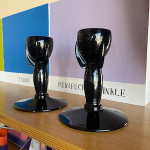 Può includere: Due portacandele in vetro nero con un design unico. I portacandele hanno una base rotonda e uno stelo alto e sottile con una parte superiore a forma di coppa. I portacandele sono su uno scaffale di legno.