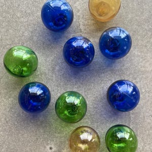Puede incluir: Un surtido de canicas de vidrio de colores azul, verde y ámbar. Las canicas son redondas y reflectantes, con un acabado brillante. Las canicas están dispersas sobre una superficie gris.