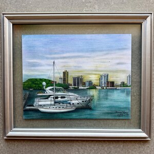 Op de afbeelding: Een ingelijst kunstwerk dat een havengezicht toont met twee jachten aangemeerd in turquoise water. De achtergrond toont een skyline onder een blauwe en gele lucht. Het kunstwerk zit in een zilveren lijst.