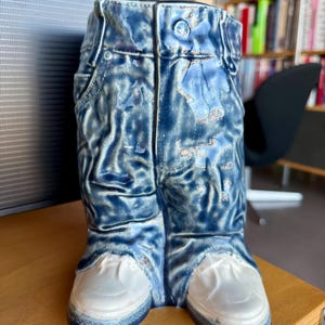 May include: A ceramic vase shaped like blue denim jeans with white sneakers. The vase has details like pockets, belt loops, and a button. The denim has a distressed look with lighter blue highlights. The sneakers are a solid white color.