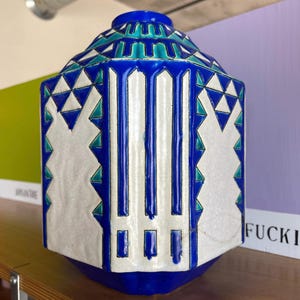 Könnte beinhalten: Geometrische Keramikvase in Blau, Weiß und Türkis. Die sechseckige Vase hat vertikale blaue Streifen und dreieckige Muster. Der Boden und der obere Rand sind tiefblau. Ein dekoratives Wohnaccessoire.