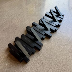 Puede incluir: Un perchero de pared negro que presenta una serie de barras rectangulares dispuestas en un patrón escalonado y superpuesto. Varias de estas barras están articuladas y pueden desplegarse hacia afuera para funcionar como ganchos.
