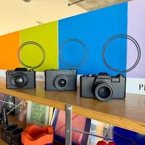 May include: Three black toy cameras are displayed on a wooden shelf. The cameras have a classic design with a lens and a rectangular body. The background features a colorful wall with orange, green, blue, and purple sections.