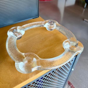 Puede incluir: Un anillo de vidrio transparente con una superficie texturizada y tres secciones redondeadas en relieve. El anillo descansa sobre una superficie de madera con un detalle de malla metálica. El diseño es moderno y minimalista.