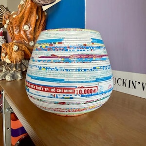 May include: A decorative, round vase made from layered paper, featuring text and colorful designs. The vase has a wide base and tapers towards the top. The text includes "SO KIEN THIET TP. HO CHI MINH 10.000d".