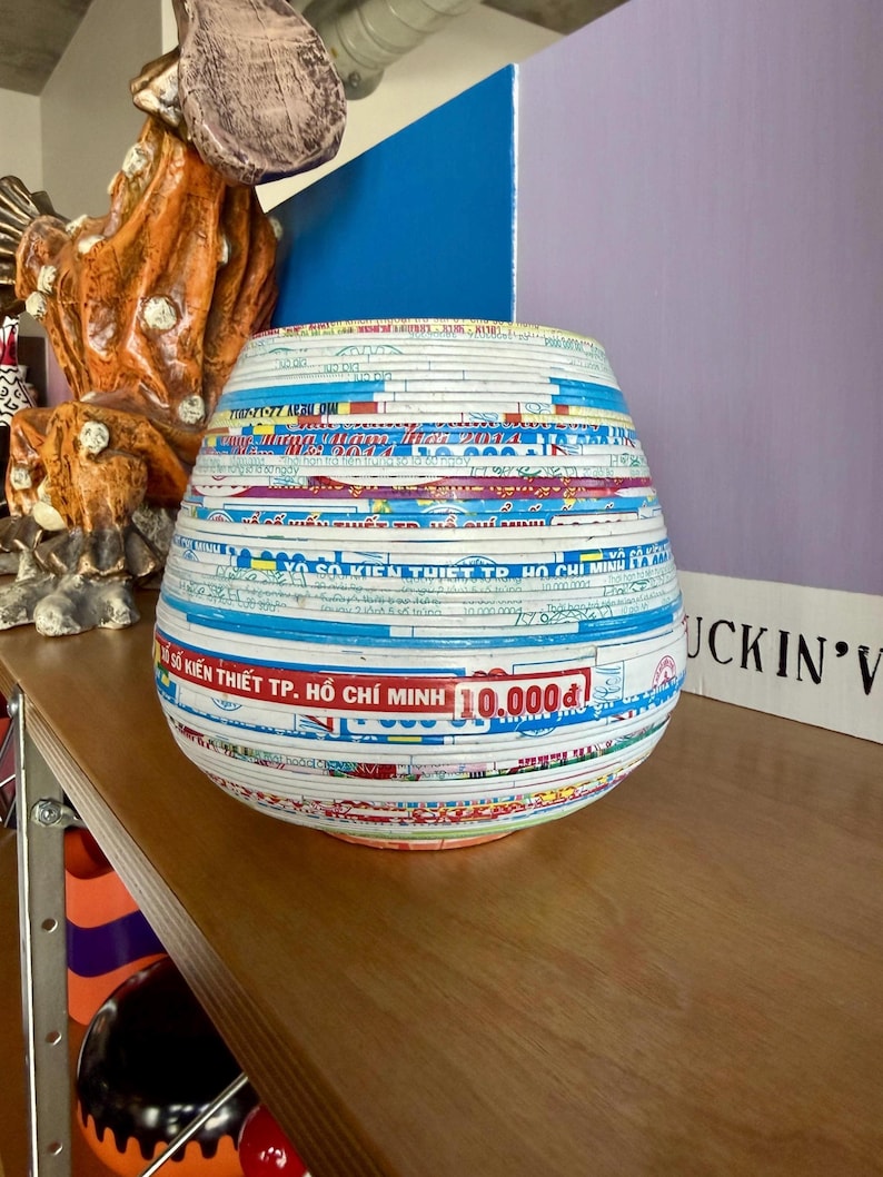 May include: A decorative, round vase made from layered paper, featuring text and colorful designs. The vase has a wide base and tapers towards the top. The text includes "SO KIEN THIET TP. HO CHI MINH 10.000d".