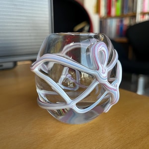 Può includere: Un vaso di vetro trasparente con un design a spirale bianco e viola. Il vaso ha una forma rotonda e un'ampia apertura.