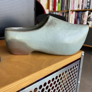 May include: A ceramic clog-shaped planter in shades of pale green and brown. The planter has a smooth, glossy finish and is designed to resemble a traditional Dutch wooden shoe. It is placed on a wooden surface.